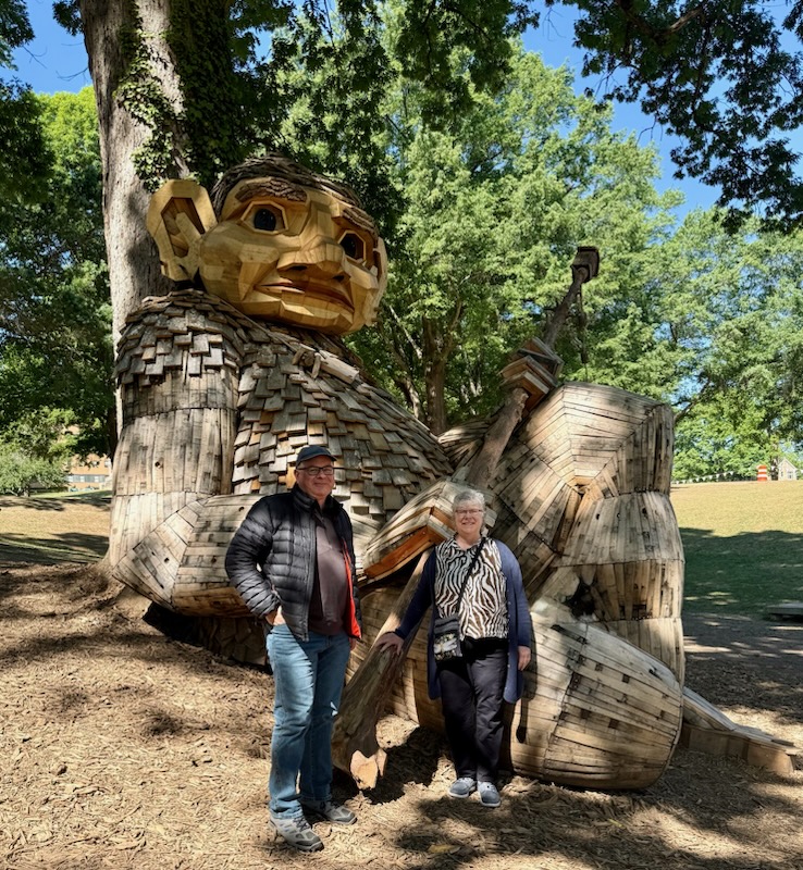 a man and woman standing in front of a wooden troll that is 5 times the size of the adults
