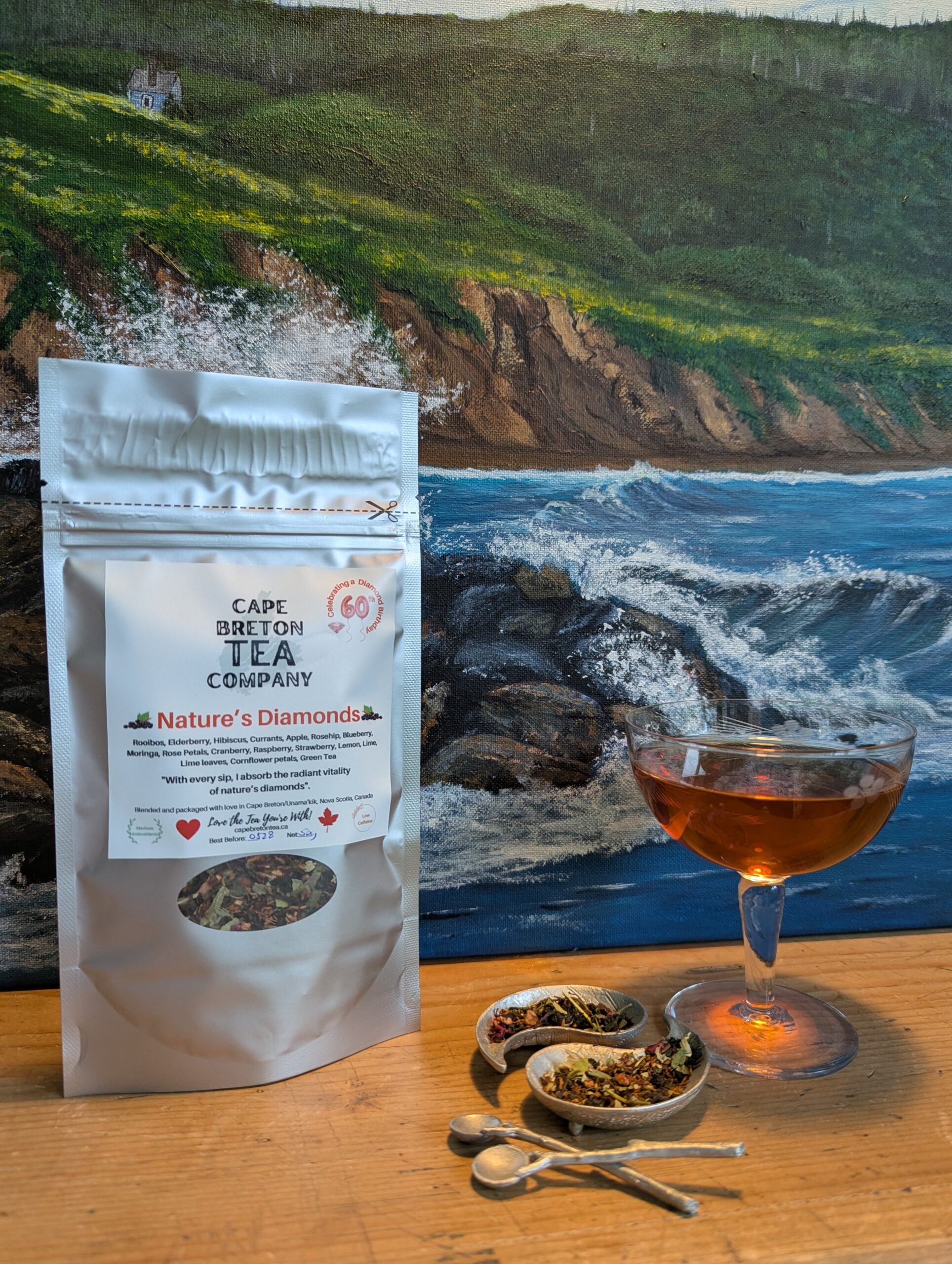 tea package, herbs in pewter with spoons, champagne glass with tea and painting of the ocean as a backdrop