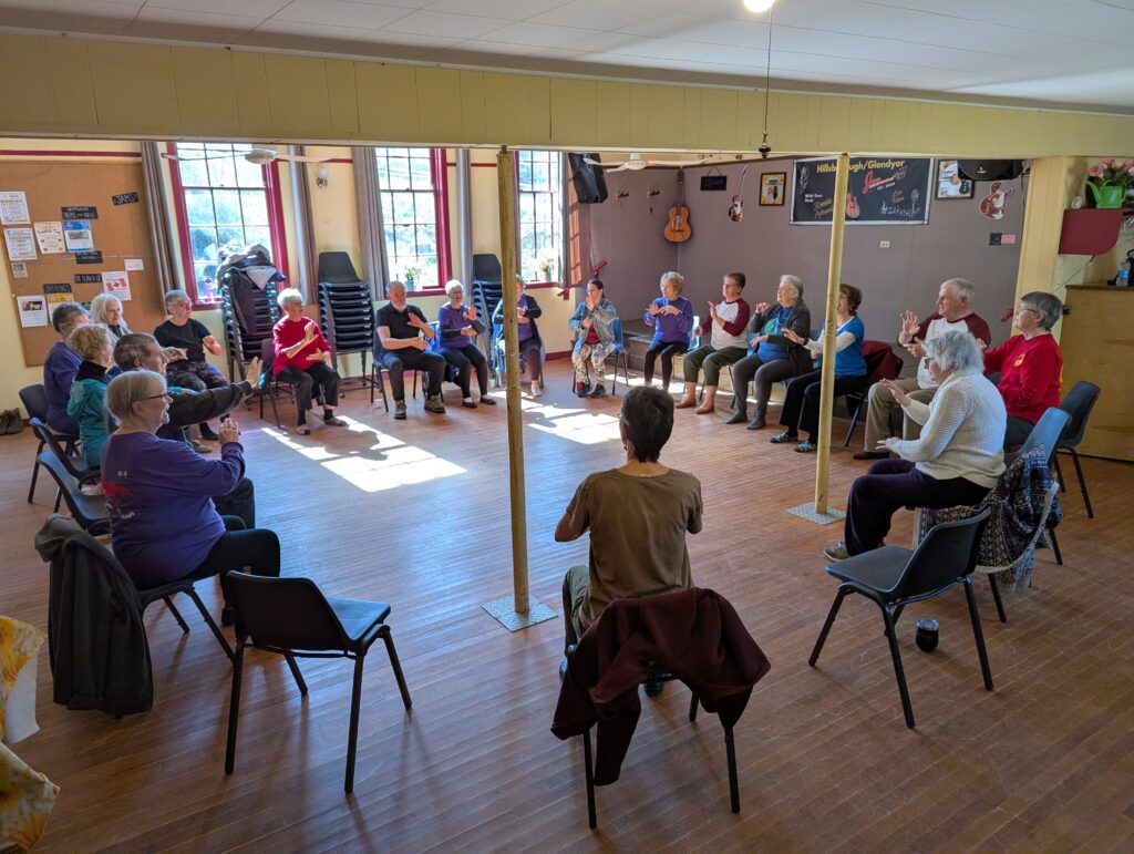 a circle of chairs in a community hall with sun shining through the windows, people all seated doing Tai Chi